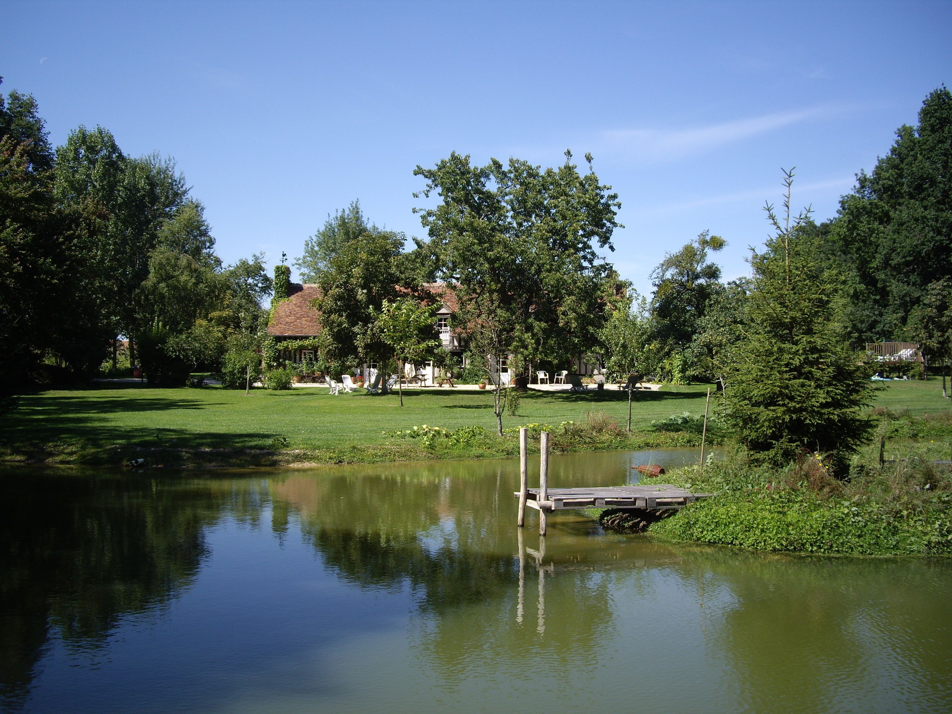 Eine friedliche Seenlandschaft mit einem hölzernen Steg, der ins Wasser führt, umgeben von üppigem Grün und einem Gebäude mit rotem Dach in der Ferne.