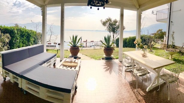 A covered outdoor seating area with a sofa and table, potted plants, and a view of the sea and mountains. - Villa Anastasye (Predore)