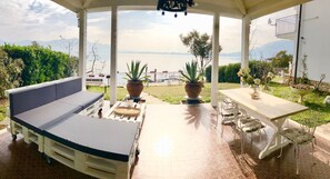 A covered outdoor seating area with a sofa and table, potted plants, and a view of the sea and mountains. - Villa Anastasye (Predore)