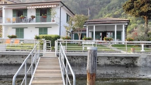 A wooden dock leading to a building with a balcony, surrounded by greenery and a mountain backdrop. - Villa Anastasye (Predore)