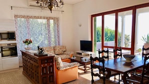 A modern kitchen with a built-in oven, a dining area with a wooden table and chairs, a sofa with floral upholstery, and a large window with a view of greenery. - Villa Anastasye (Predore)