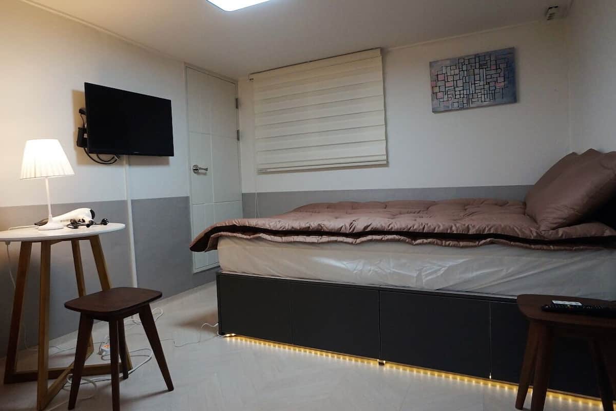 Ein gemütliches Schlafzimmer mit Bett, Schreibtisch, Stuhl, Fernseher und einem Fenster mit Jalousien.