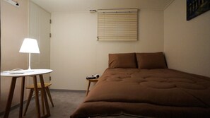 A bedroom with a bed, a bedside table, a lamp, and a window with blinds. - Blue Mansion Guesthouse (Seoul)