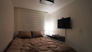 A small, dimly lit room with a single bed, a mounted TV, and a bedside table with a lamp. - Blue Mansion Guesthouse (Seoul)