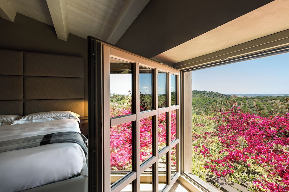 A bedroom with a large window, a bed, and a view of pink flowers outside.