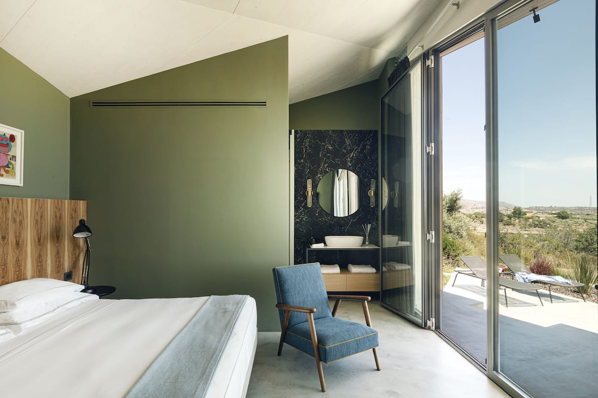 A bedroom with a large bed, a chair, a vanity with a mirror, and a view of the landscape through a sliding door.