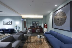 A modern living room with a blue sofa, grey armchairs, and a coffee table. There is a kitchen area in the background with a bar and seating. - M Suite Hotel (Algiers)