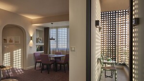 A dining area with a round table and chairs, a sofa, and a small table with a lamp. There is a window with curtains and a wall-mounted shelf with decorative items. - M Suite Hotel (Algiers)