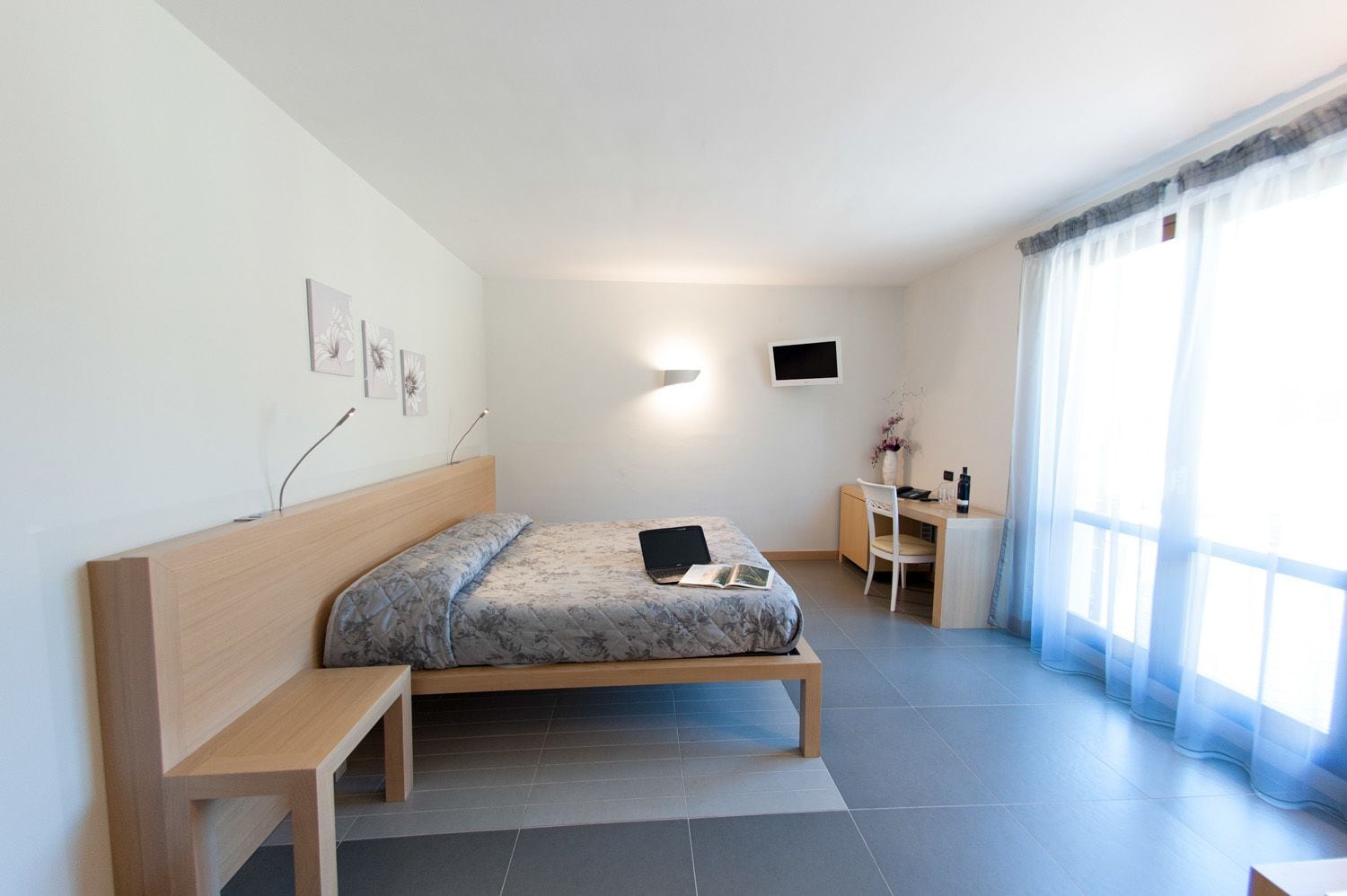 A modern bedroom with a bed, bedside table, desk, chair, and a TV on the wall.