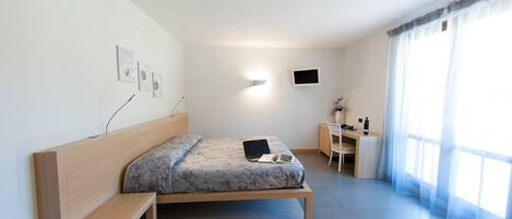 A modern bedroom with a bed, bedside table, desk, chair, and a TV on the wall.