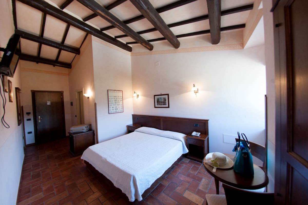 A hotel room with a large bed, a wooden headboard, a nightstand, a desk with a chair, and a small table with a vase.