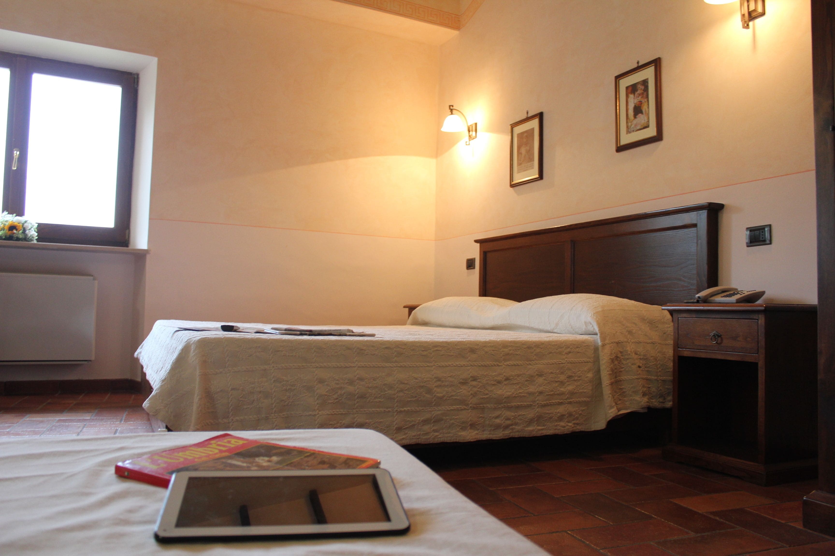 A bedroom with a bed, bedside table, nightstand, lamp, and a window with a view.