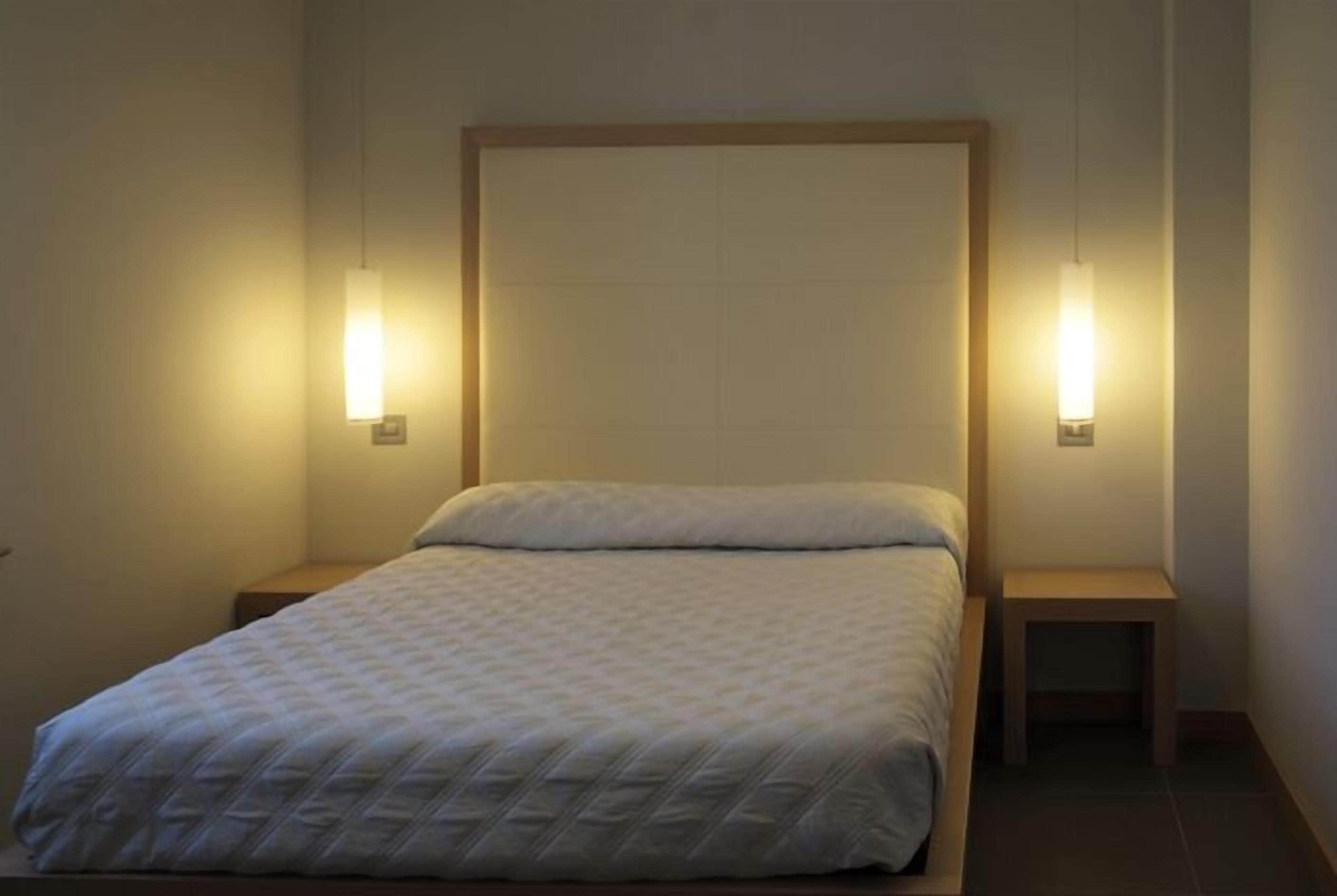 A neatly made bed with a quilted blanket in a simple room with two bedside tables and wall-mounted lights.