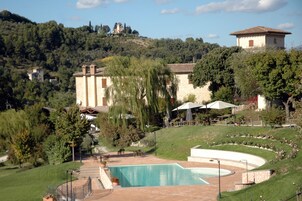Zona de piscina con solárium de piedra, rodeada de vegetación y una colina con edificios al fondo.