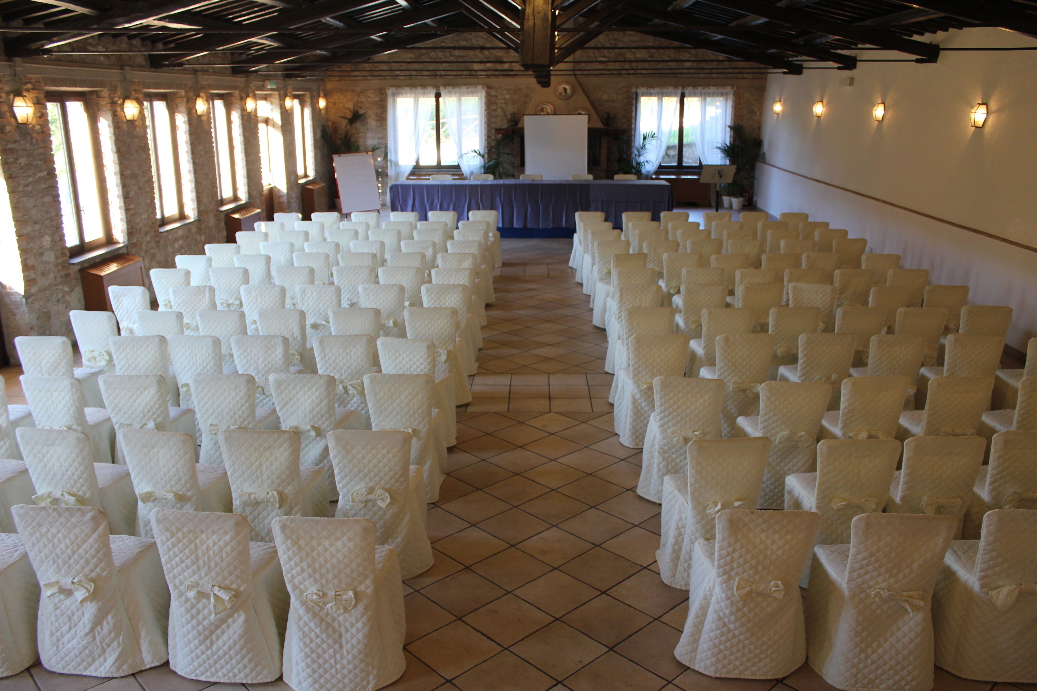 A large hall with rows of white chairs, a stage with a table, and large windows.