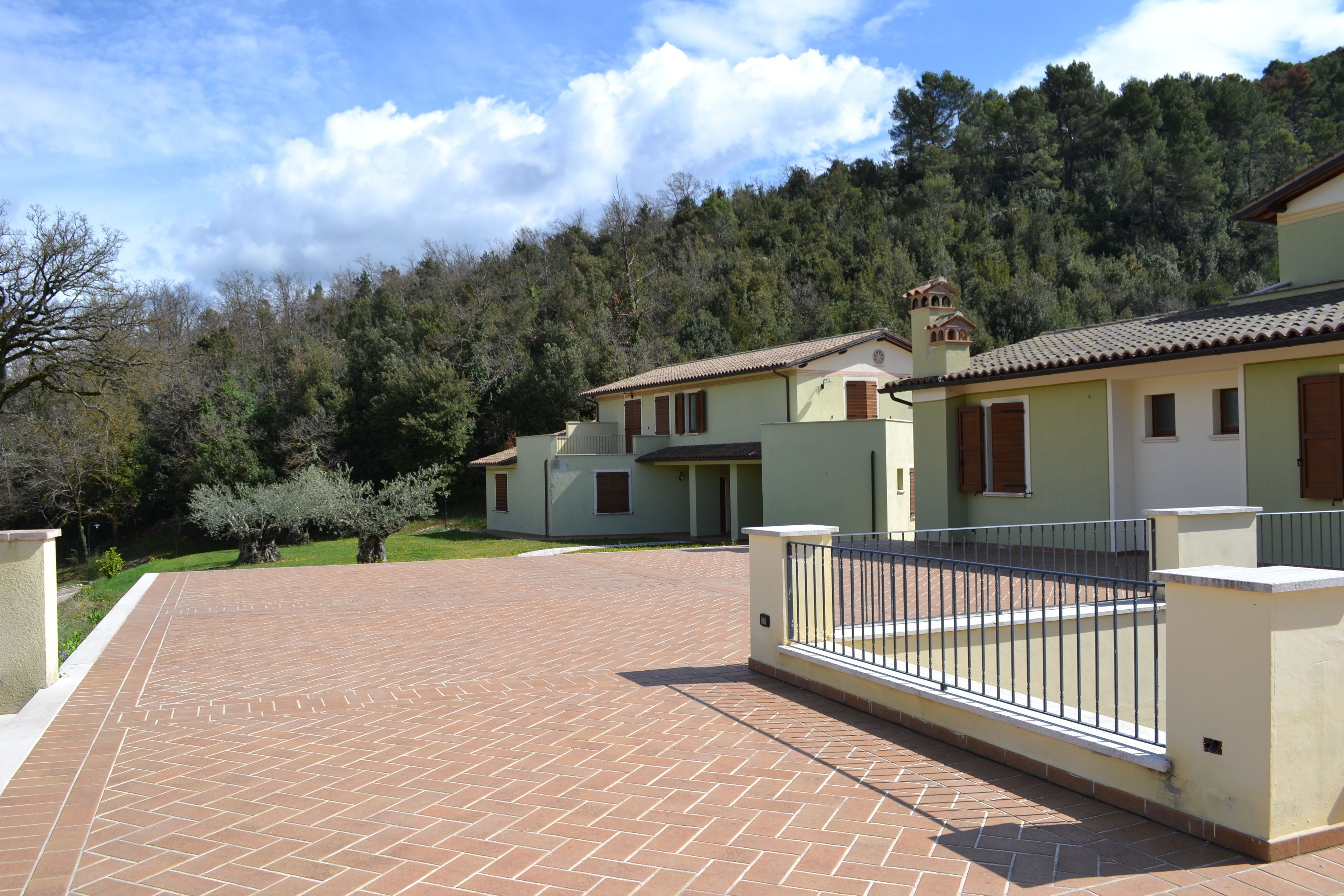 A residential building with a paved driveway, a metal fence, and a forested hillside in the background.