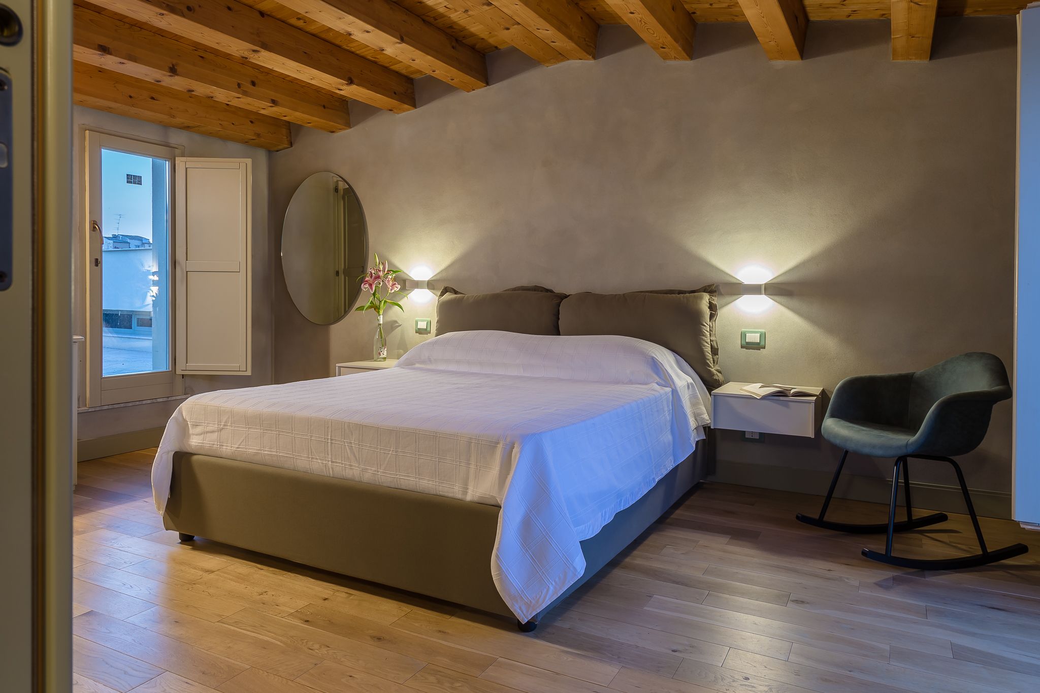 A bedroom with a bed, bedside tables, a chair, a mirror, and a view of the outside.