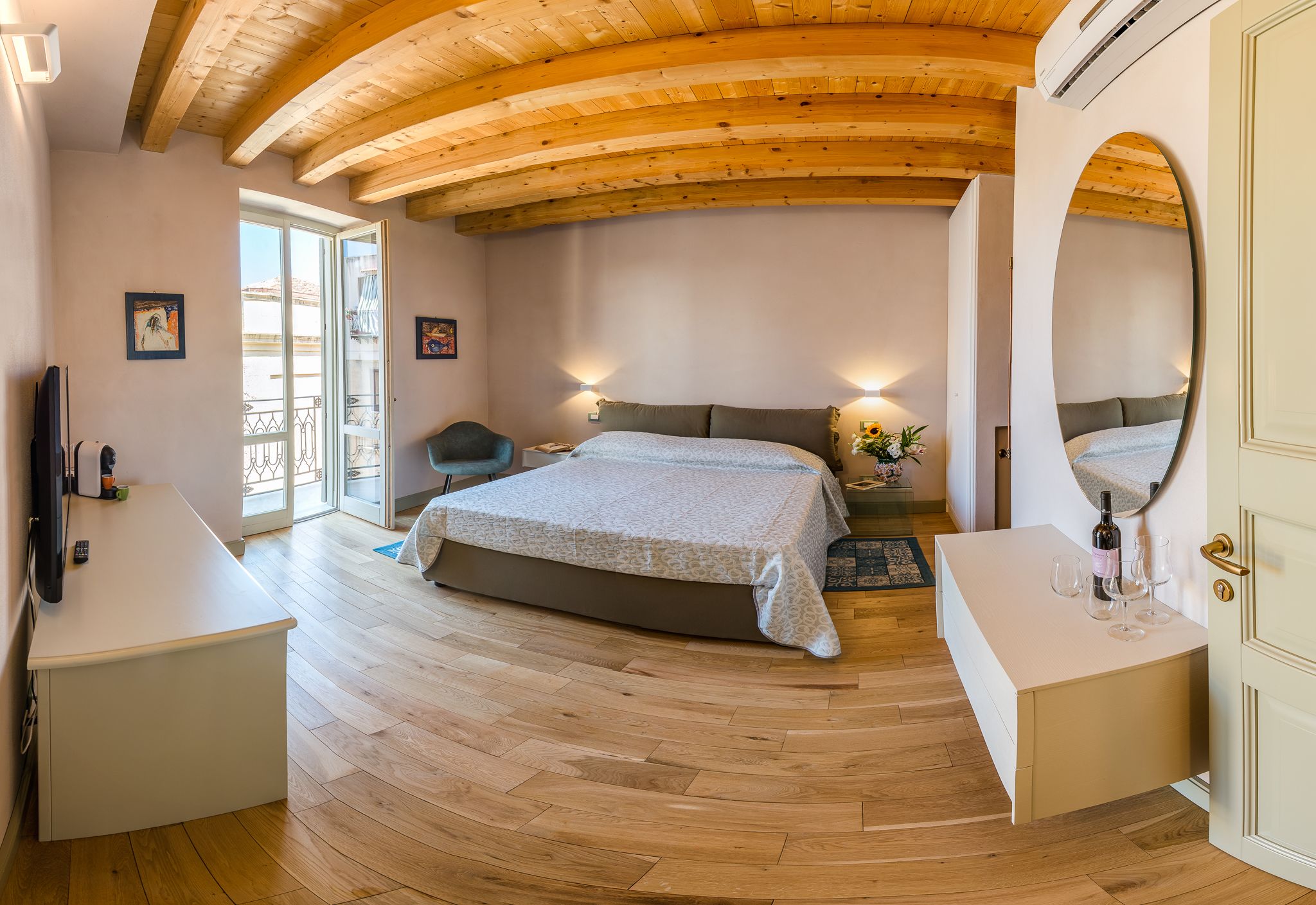 Een slaapkamer met een houten plafond, een bed met een beddegoed met een patroon, een nachtkastje met een fles en glazen, en een grote spiegel aan de muur.