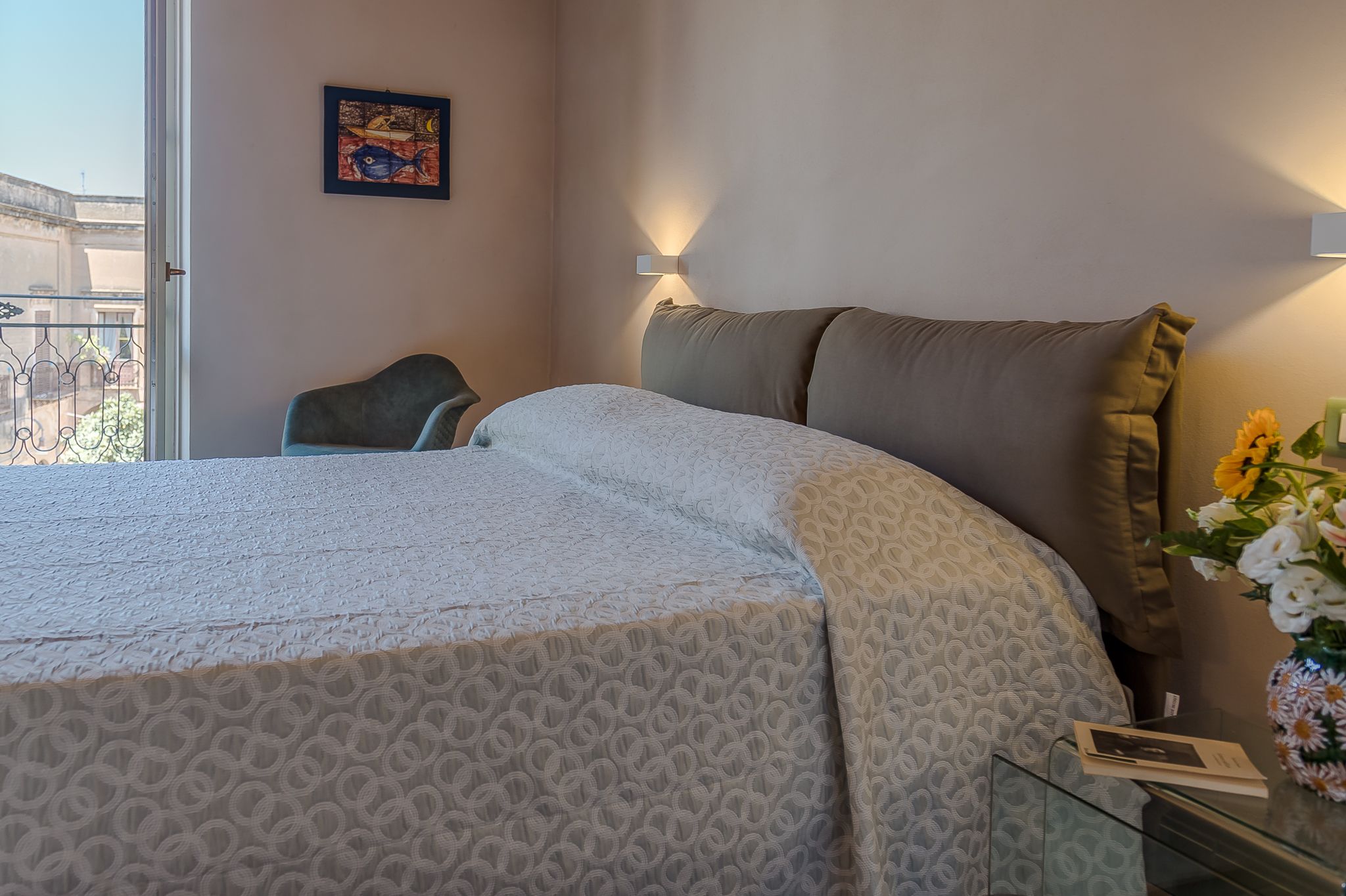 A neatly made bed with a patterned bedspread, a bedside table with a vase of flowers, and a wall-mounted light fixture.