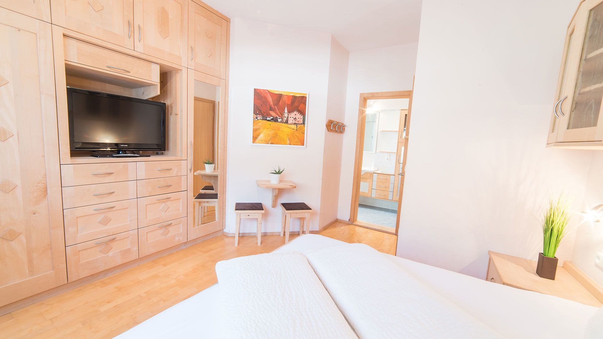 Un dormitorio con televisión, armarios de madera, un escritorio pequeño, dos sillas, una cama, un espejo y un cuadro en la pared.