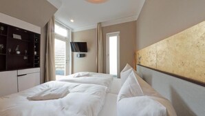 A hotel room with two beds, a TV mounted on the wall, a minibar, and a large window. - Ringelnatz Warnemünde (Rostock)