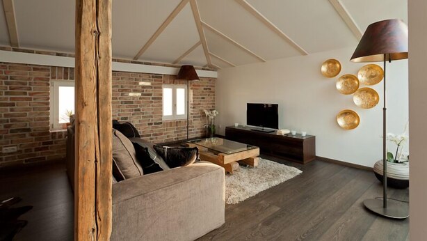 A modern living room with exposed brick walls, a wooden ceiling beam, a large sofa, a coffee table, a TV, and a floor lamp. - Ringelnatz Warnemünde (Rostock)