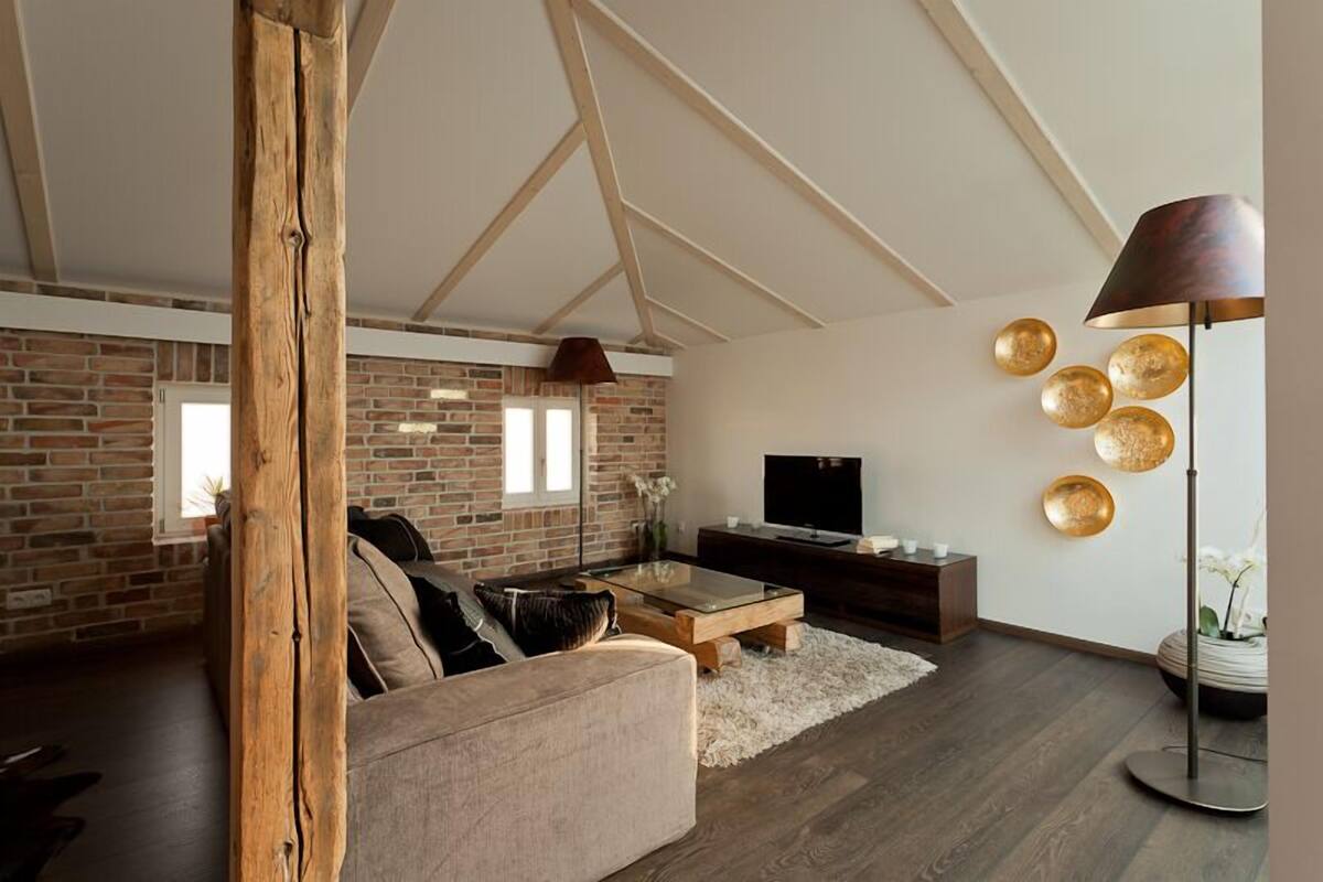 A modern living room with exposed brick walls, a wooden ceiling beam, a large sofa, a coffee table, a TV, and a floor lamp.