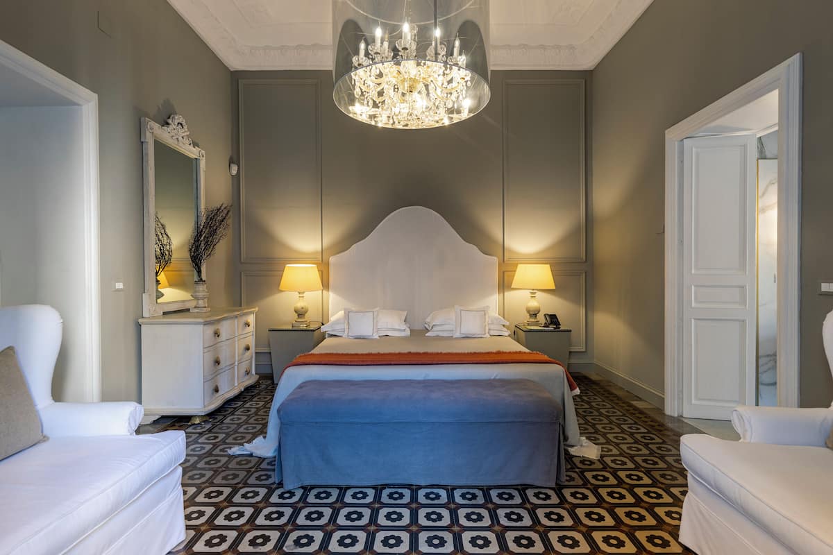 Een hotelkamer met een groot bed, twee nachtkastjes met lampen, een kroonluchter, een spiegel en vloerbekleding met een patroon.