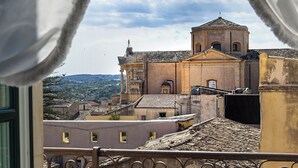 A balcony with a wrought iron railing, a table with a plate, and a chair. In the background, there is a building with a dome and a bell tower.