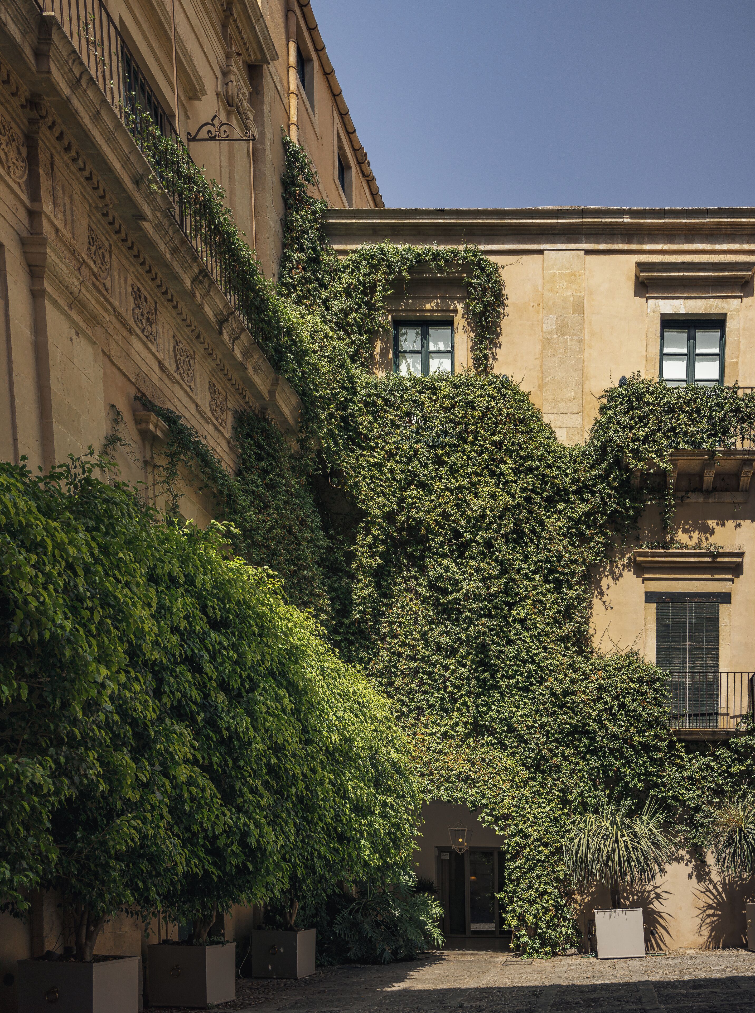 Un edificio con un cortile interno, caratterizzato da un ingresso coperto, una piccola area giardino con piante in vaso e una scala che conduce a un piano superiore.