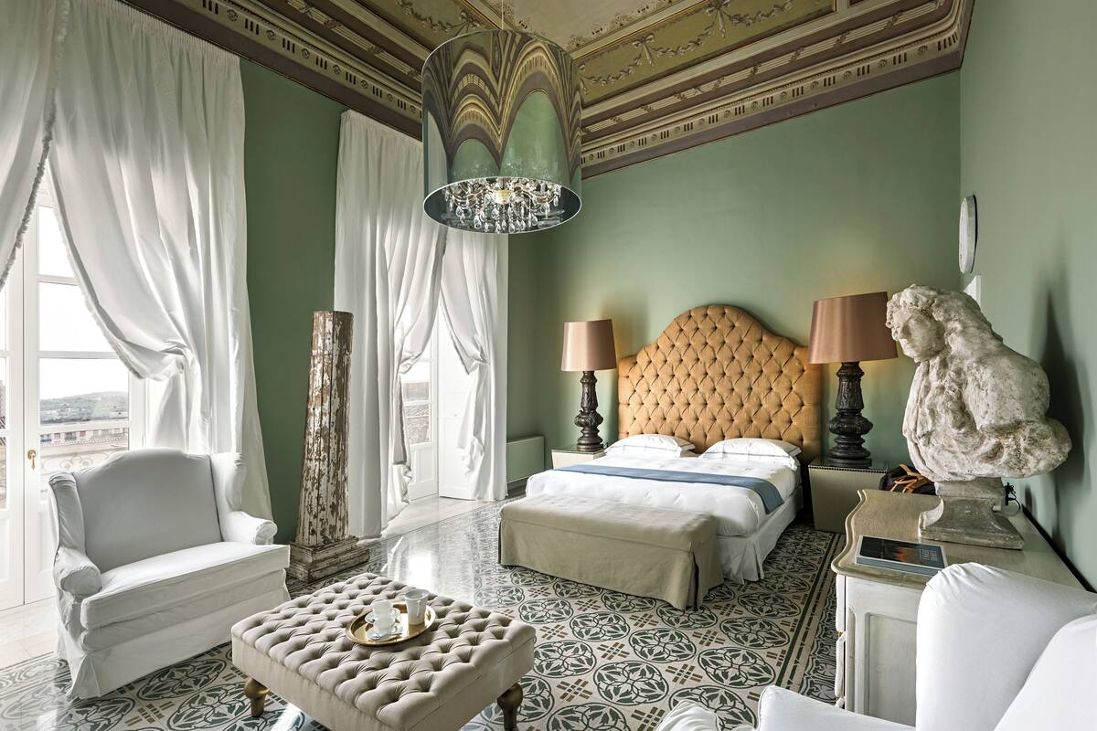 Een hotelkamer met een bed, twee nachtkastjes, een stoel, een lamp, een beeld en uitzicht op de stad.