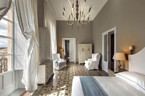 Een hotelkamer met een groot bed, twee witte fauteuils, een kroonluchter en uitzicht op het landschap.