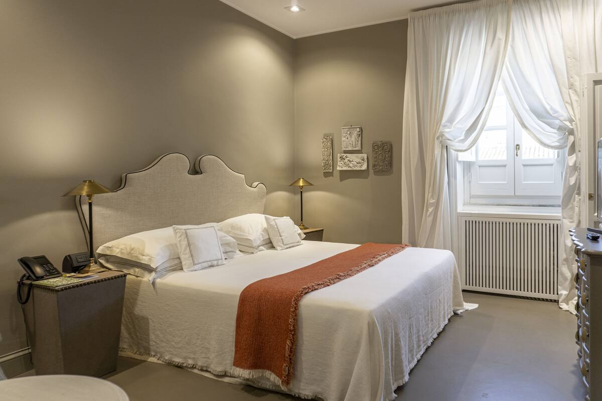 Een keurig opgemaakt bed met witte lakens en een bruine plaid, een bedlampje, een telefoon op een nachtkastje, en een raam met dunne gordijnen.