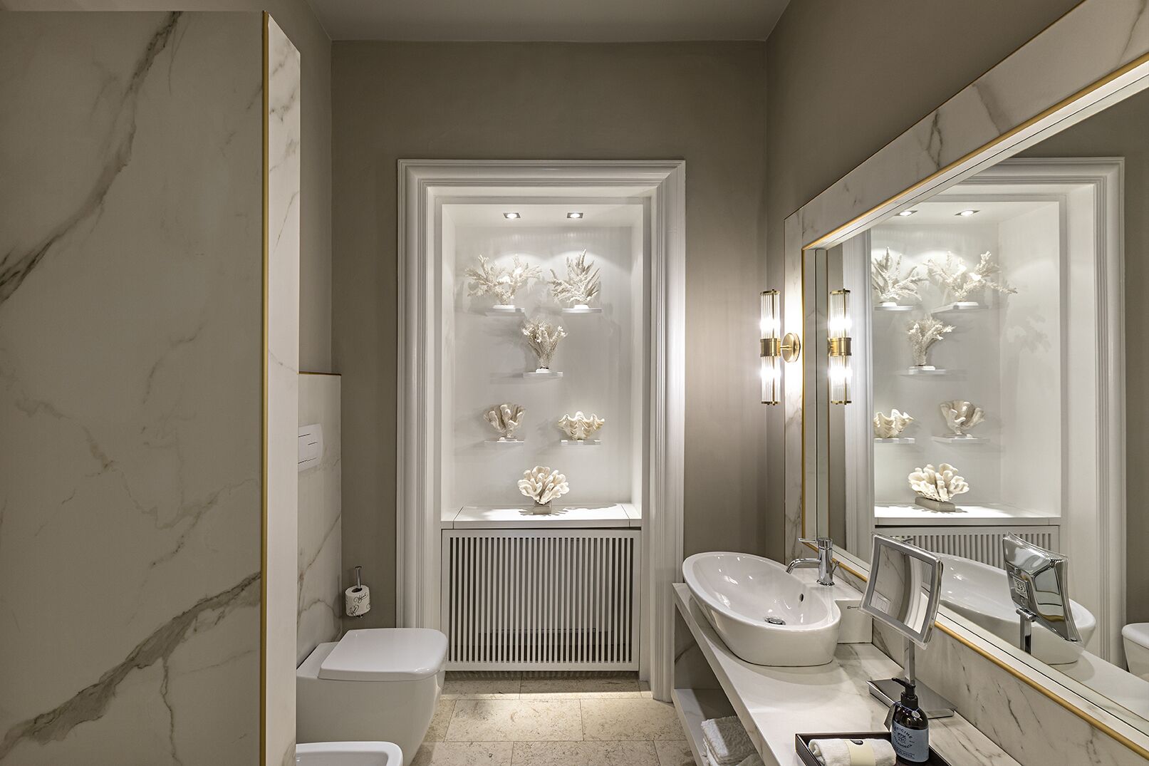 A modern bathroom with a marble floor, a white toilet, a sink with a large mirror, and a display of decorative items in the shower area.