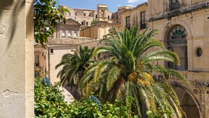 A courtyard with palm trees, a stone wall, and historic buildings with arched windows and balconies.