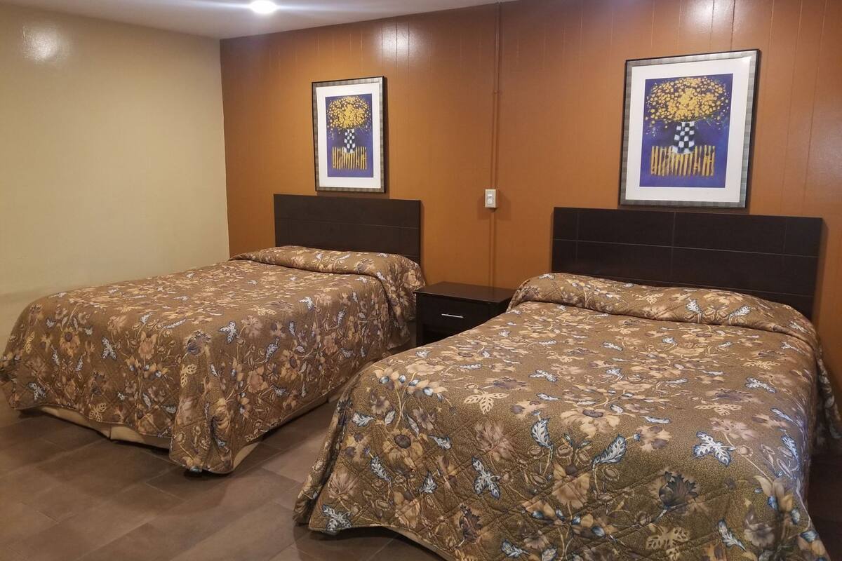 A hotel room with two beds, a dark headboard, a small bedside table, and two framed pictures on the wall.