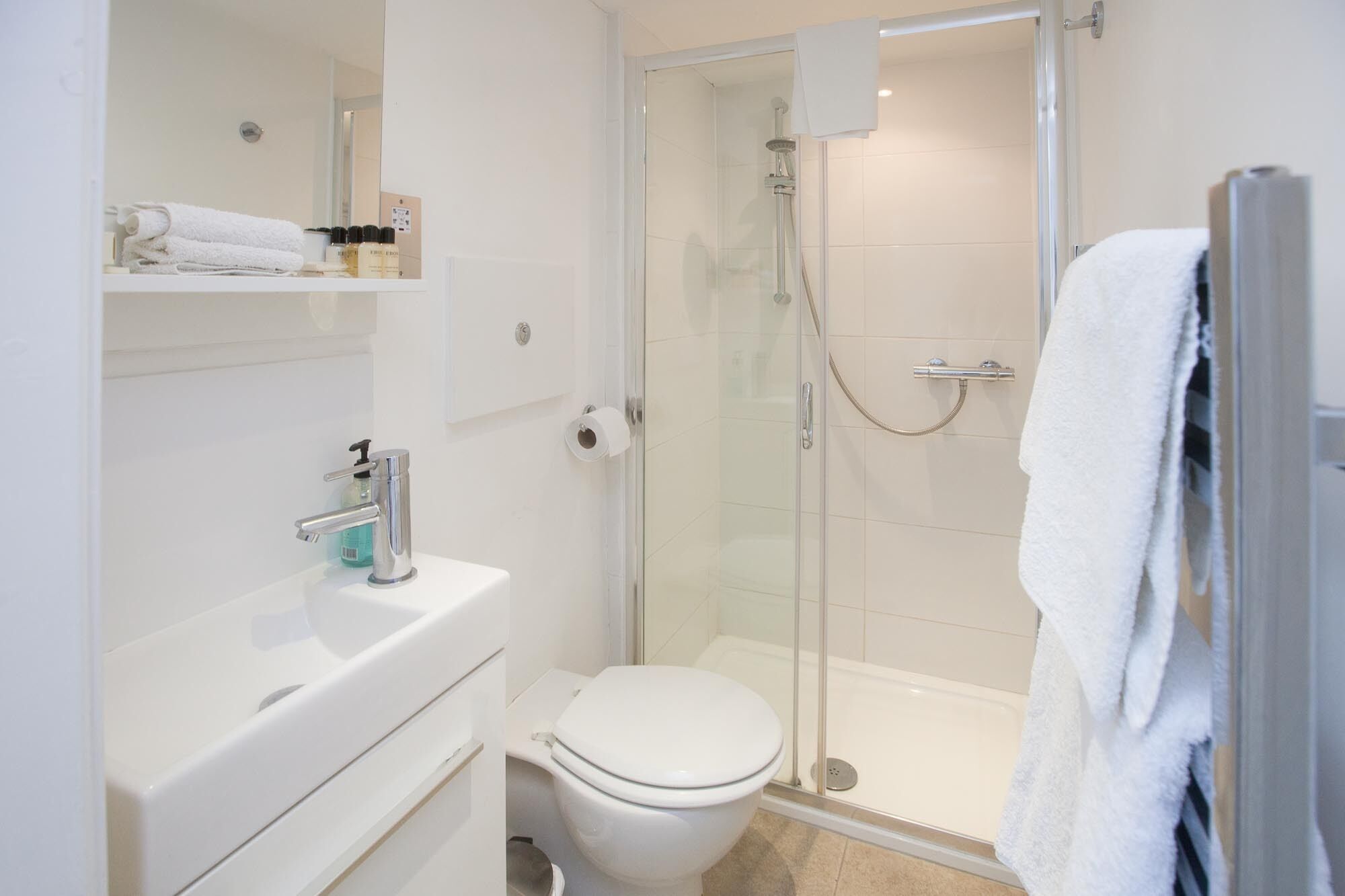 Et moderne badeværelse med en brusekabine i glas, et hvidt toilet og en vask med håndvask og sæbedispenser.