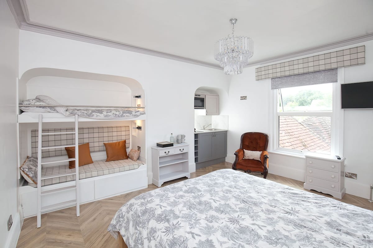 A bedroom with a bunk bed, a large bed, a chair, a desk, a mirror, and a window with a view of the outside.