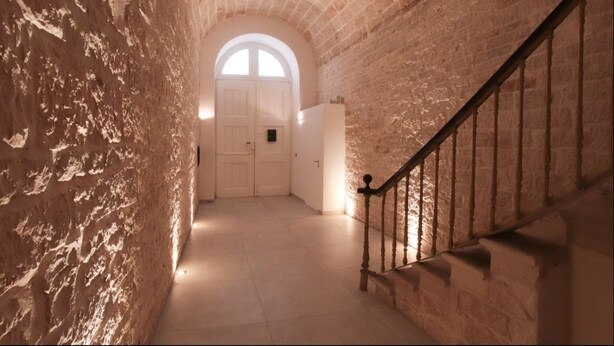 A narrow hallway with a stone wall, a wooden staircase with a metal handrail, and a white door at the end. - B&B il Castello (Giovinazzo)