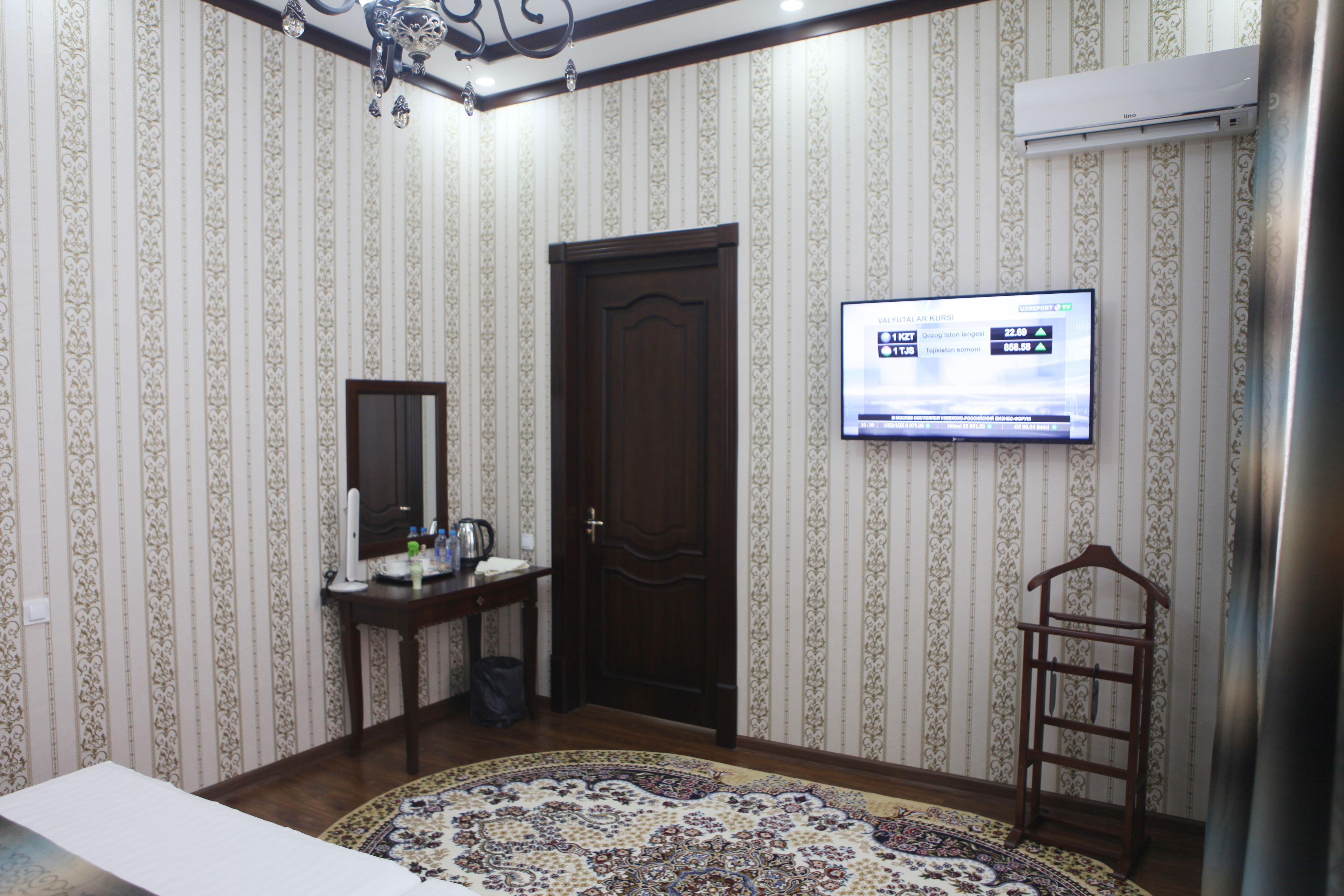 Habitación de hotel con puerta de madera, televisor montado en la pared, una mesita con tetera y una alfombra estampada.