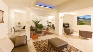 A spacious living area with a sofa, ottoman, and a dining table with chairs. There are potted plants and a television. - Hotel Corallo (Orbetello)