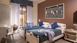 A hotel room with two beds, a desk, a chair, a bedside table with a lamp, and a painting on the wall. - Hotel Corallo (Orbetello)