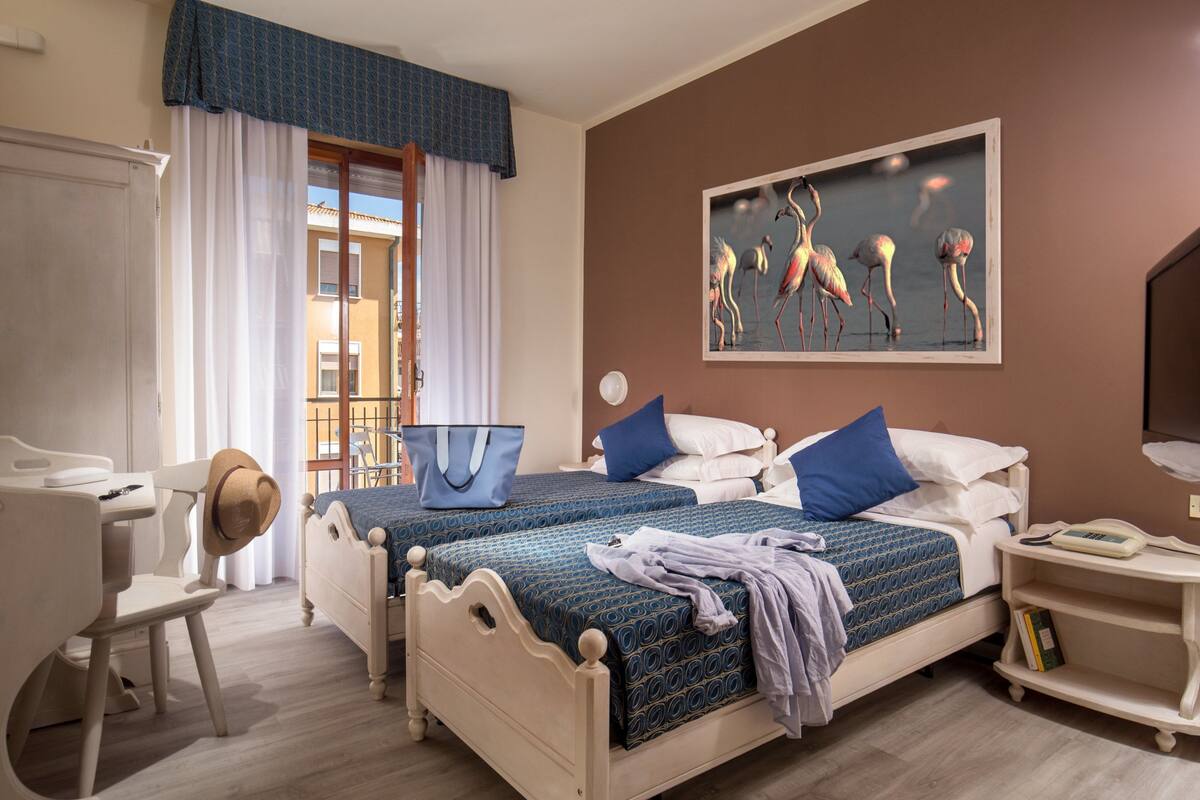 Habitación de hotel con dos camas, un escritorio, una silla, una mesita de noche con lámpara y un cuadro en la pared.