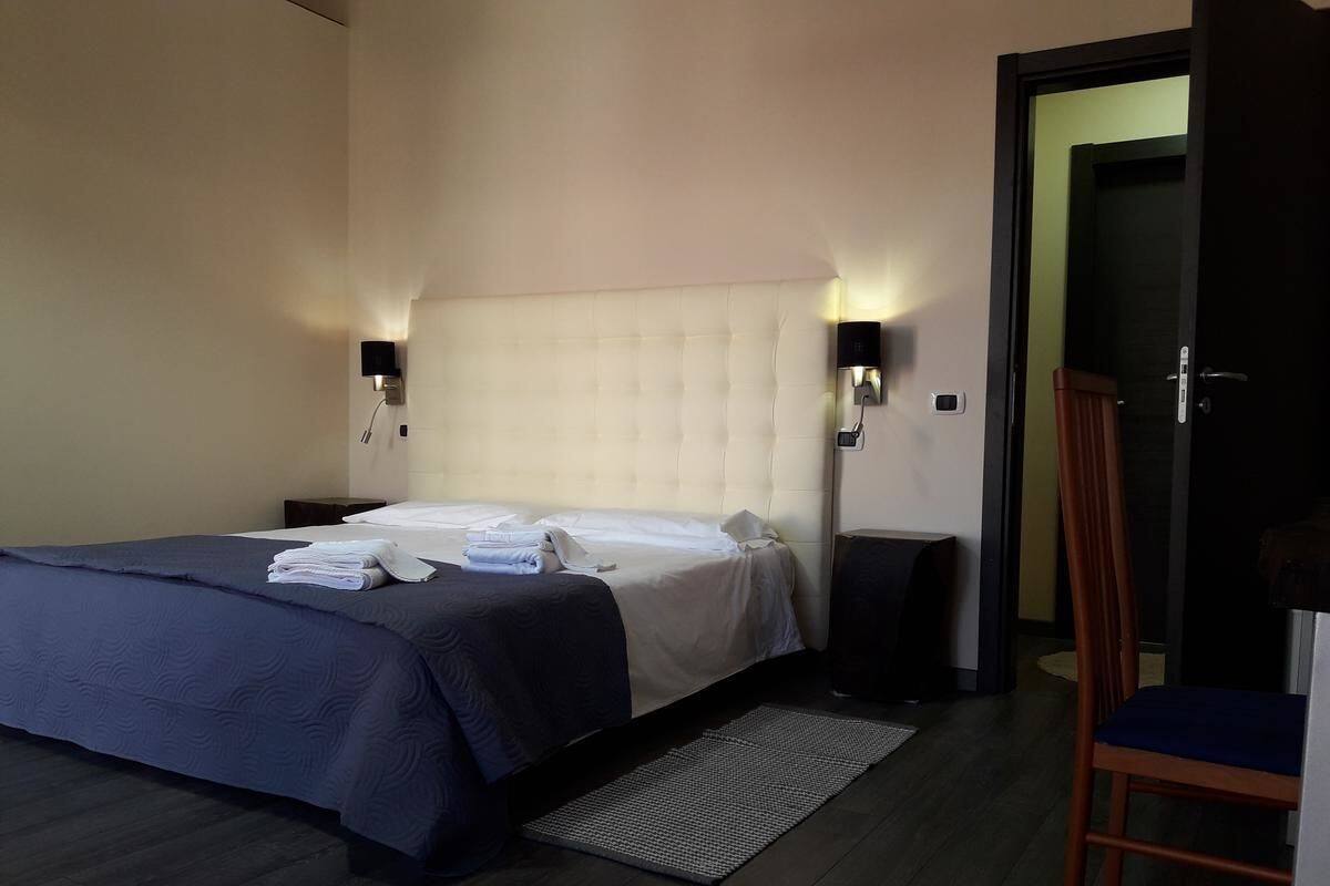 A hotel room with a bed, two wall-mounted speakers, a chair, and a door leading to another room.