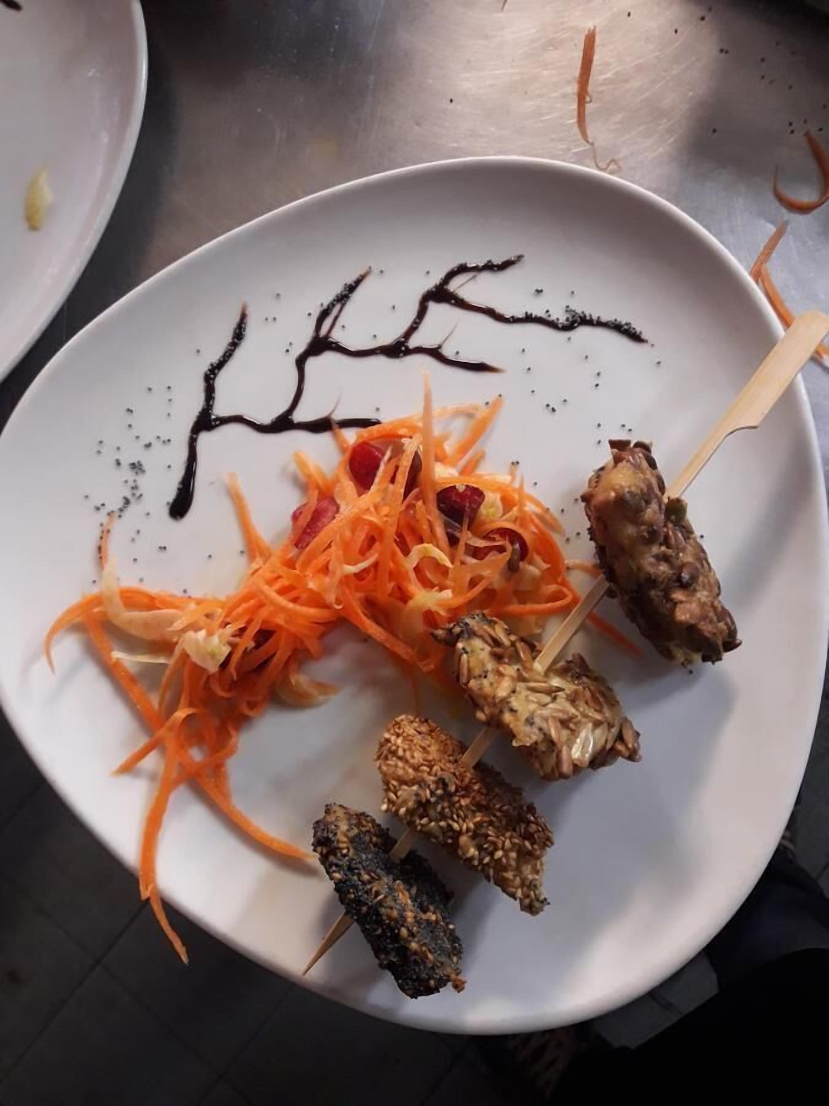 A white plate with skewered food items, including carrots, meat, and possibly nuts, on a stainless steel surface.