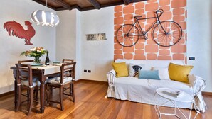 Een eethoek met houten stoelen en een tafel, een bank met kussens, een aan de muur bevestigde fiets en een muur met een decoratie van een haan.