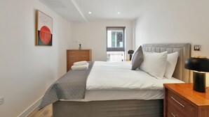 A modern bedroom with a large bed, a wooden dresser, a bedside table with a lamp, and a window with blinds. - 1Bed & 2Beds Apartment in Liverpool St by City Stay Aparts London (London)