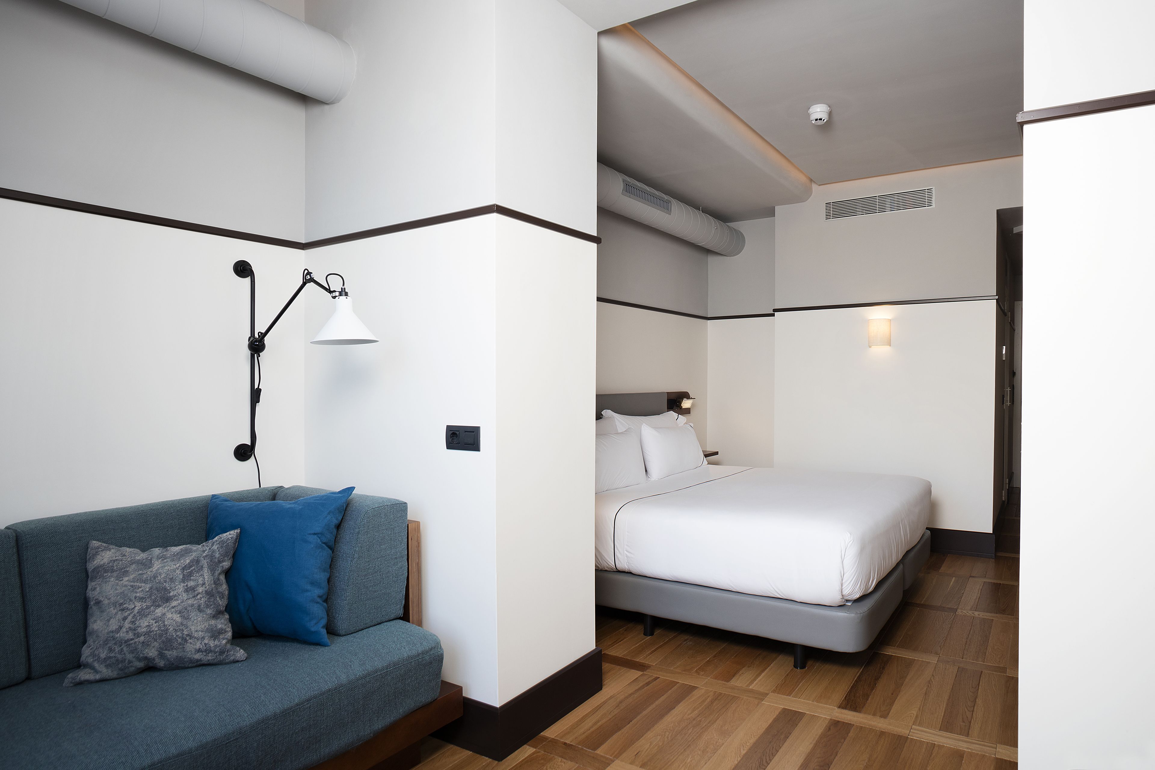 Una habitación de hotel moderna con un sofá gris, una cama y una mesita de noche con una lámpara.