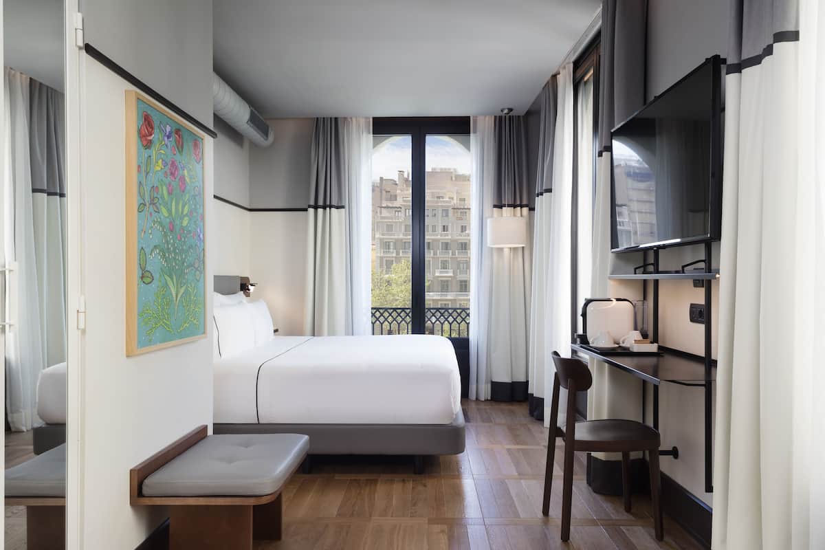 A hotel room with a bed, a desk, a chair, a TV, and a balcony with a view of buildings.