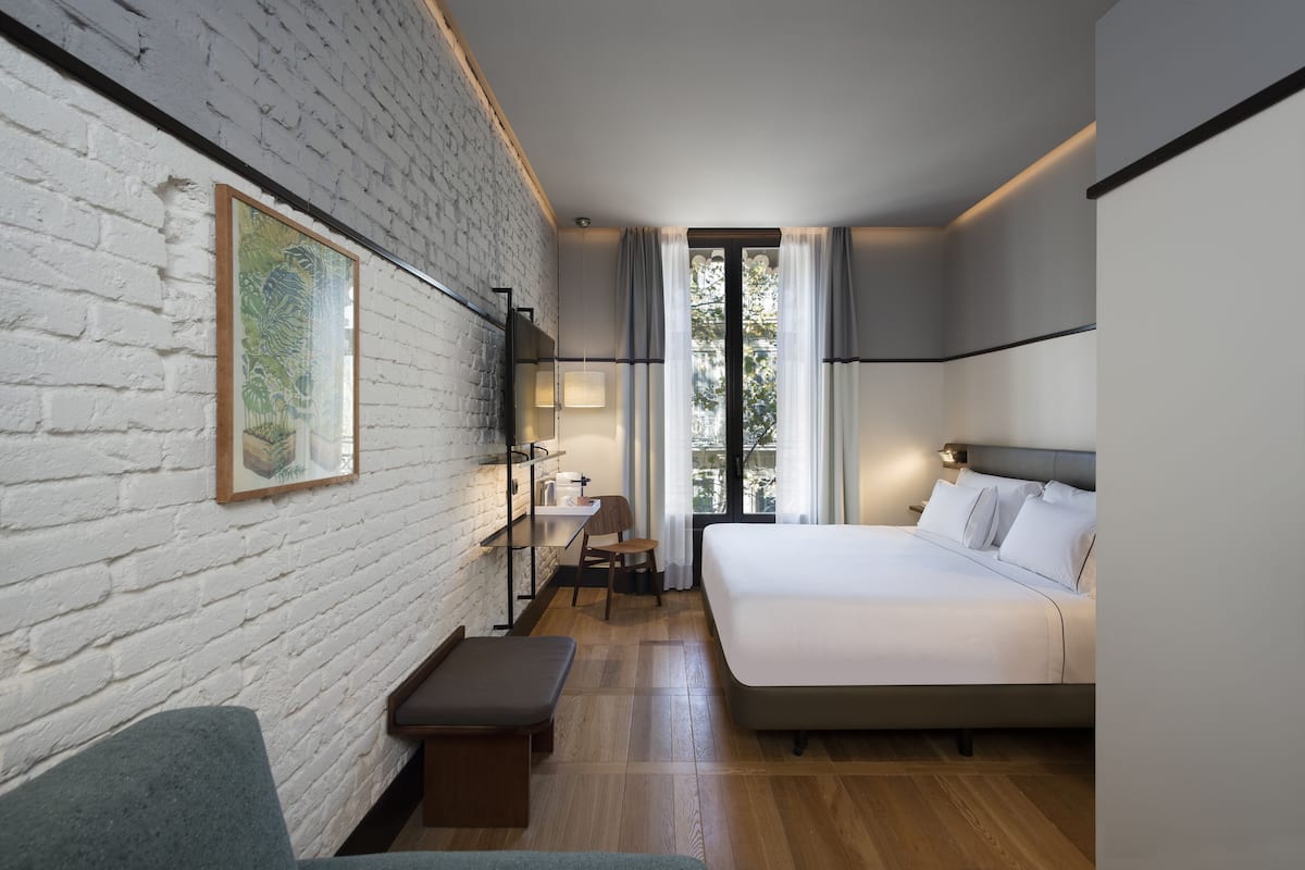 A modern hotel room with a white brick wall, a large bed, a desk with a chair, and a window with curtains.